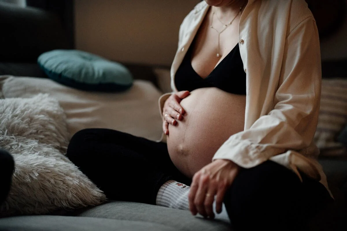 Mutter sitzt mit Babybauch auf dem Sofa und haelt die Hand darauf