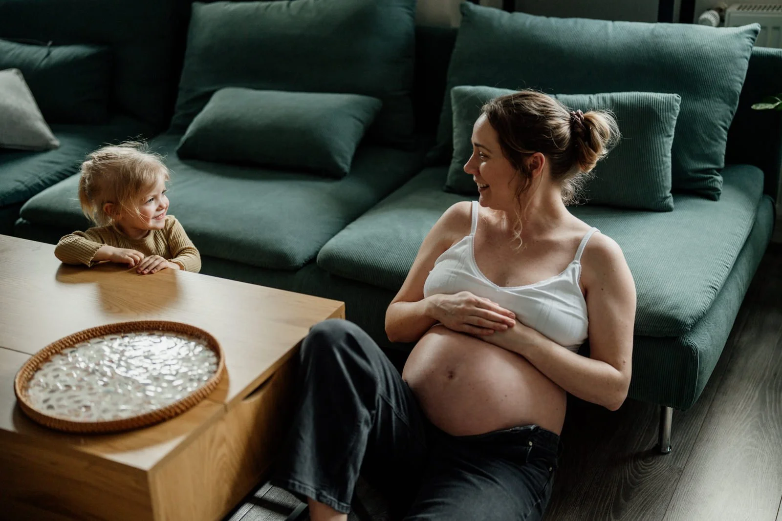 schwangere mama sitzend ans sofa gelehnt schaut lachend zu kleinkind