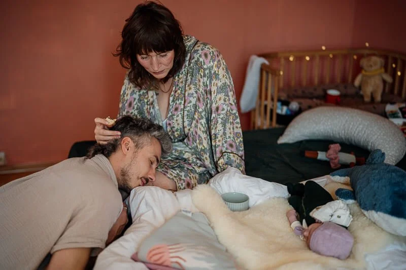 Erschöpfte Eltern auf Bett sitzend während Baby neben ihnen liegt