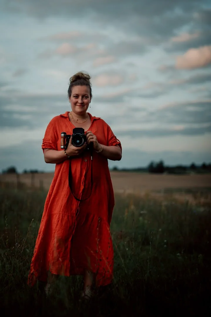 Frau in einem roten Kleid steht auf Wiese mit wolkigem Himmel im Hintergrund, hält eine große analoge kamera in der hand und lächelt in die kamera.