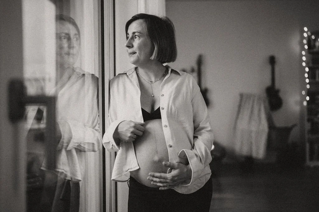 Schwangere Frau steht nachdenklich am Fenster Schwarzweiß, emotionale Schwangerschaftsfotografie