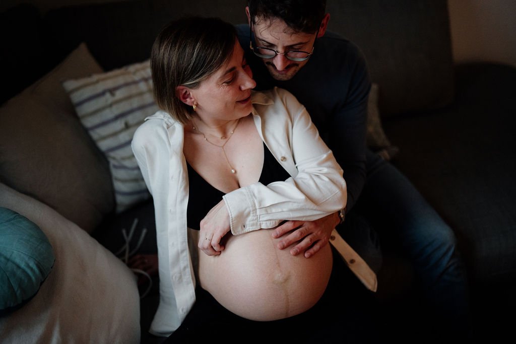 Innige Umarmung schwangeres Paar auf Sofa, emotionale Schwangerschaftsfotografie zuhause