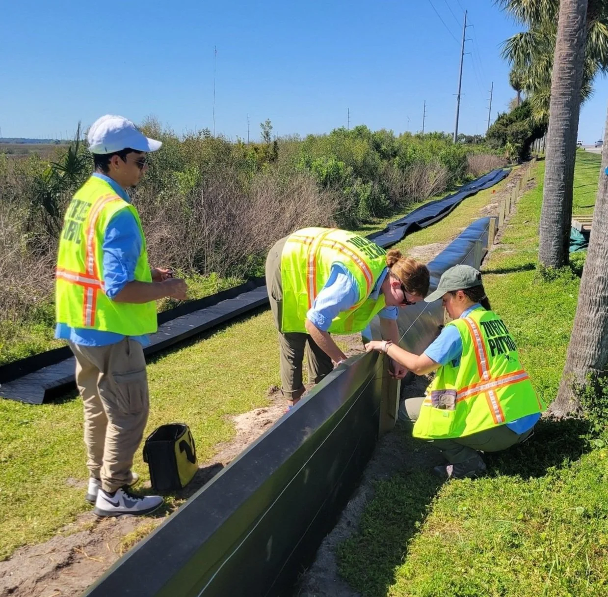 Three people wearing yellow vests labeled 'Utility Patrol' working outdoors on a sunny day, installing Animex Fencing along a grassy area with trees and shrubbery nearby.