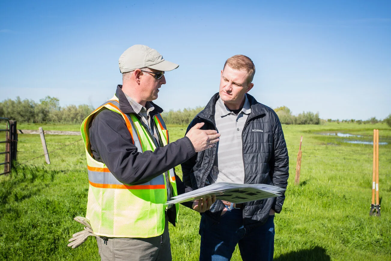 Two men outdoors in a grassy field, one wearing a safety vest and hat, explaining something to the other who is holding a clipboard, with shovels and a fence in the background.