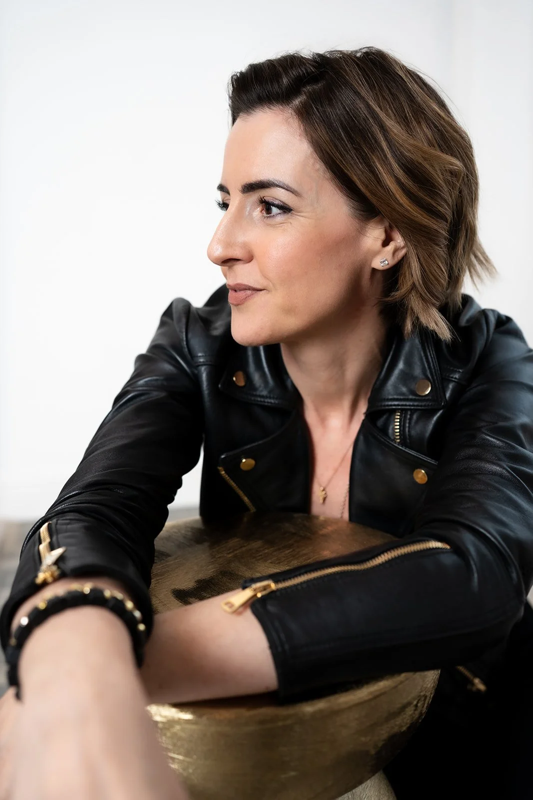 Lisa Moore, Sovereign Frequency Architect, in a black leather jacket leaning contemplatively on a gold stool, gazing with quiet sovereign authority