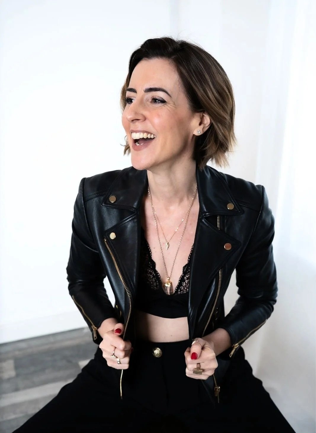 Lisa Moore, founder of House of Soul Sovereignty, wearing a black leather jacket, black lace top, and gold jewelry, smiling and holding the lapels of her jacket.