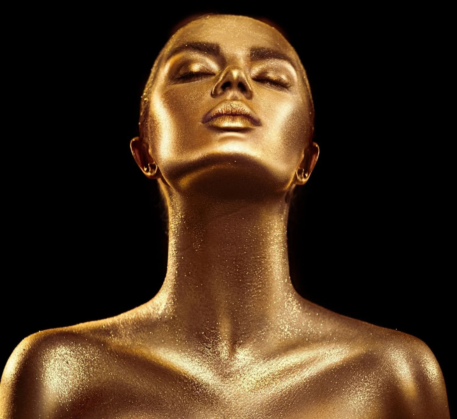 Portrait of a woman covered in gold body paint with closed eyes and a black background.
