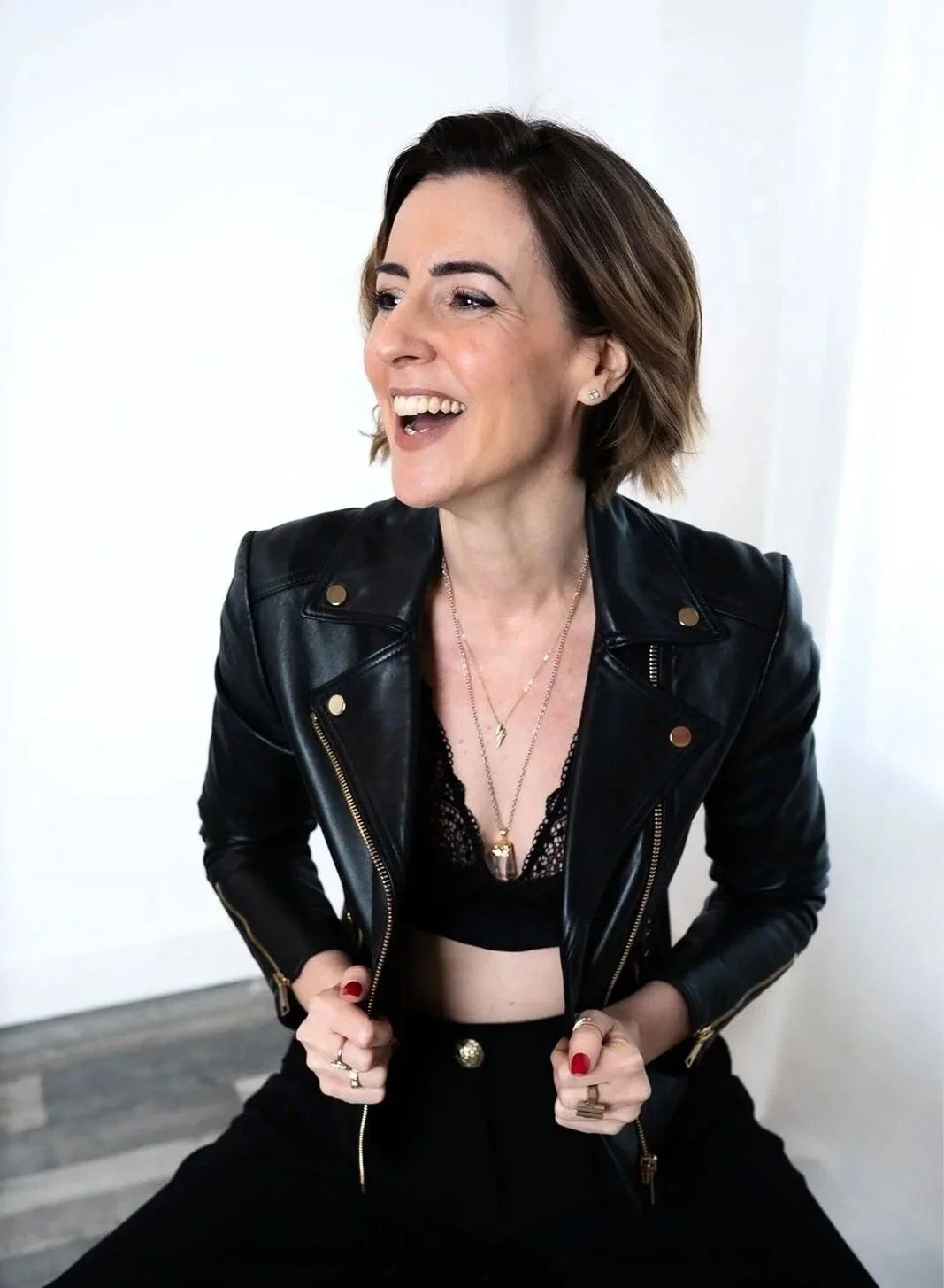 Lisa Moore, founder of House of Soul Sovereignty, wearing a black leather jacket, black lace top, and gold jewellery, smiling and holding the lapels of her jacket.