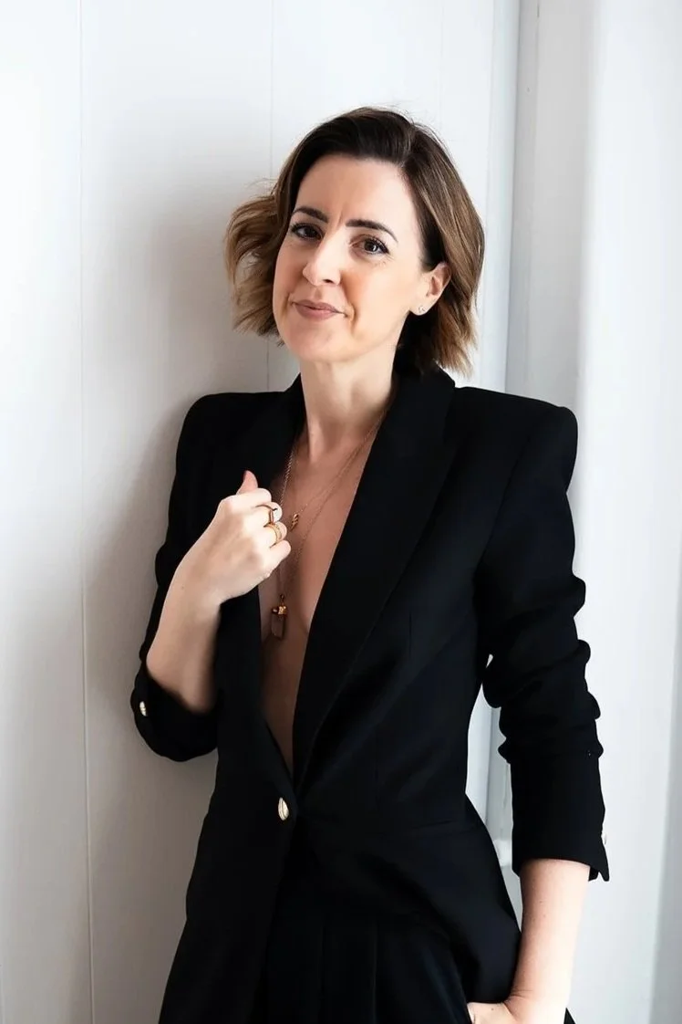Lisa Moore in black blazer with jewellery, against white wall — Sovereign Frequency Architect + Speaker — House of Soul Sovereignty