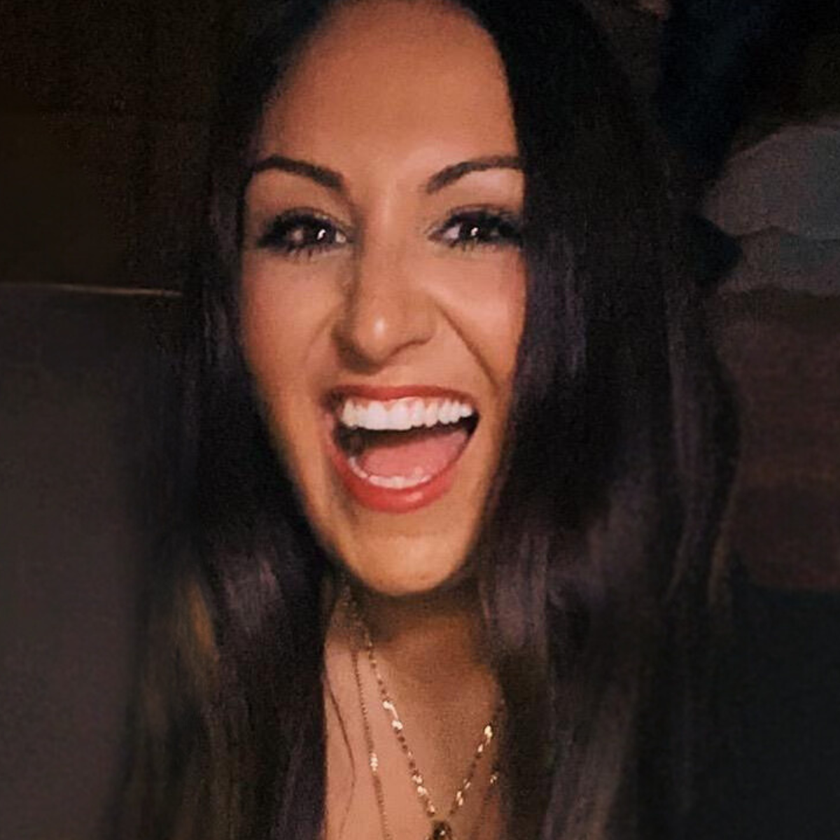 A woman with long, brown hair smiling at the camera, wearing a black top and gold necklaces.