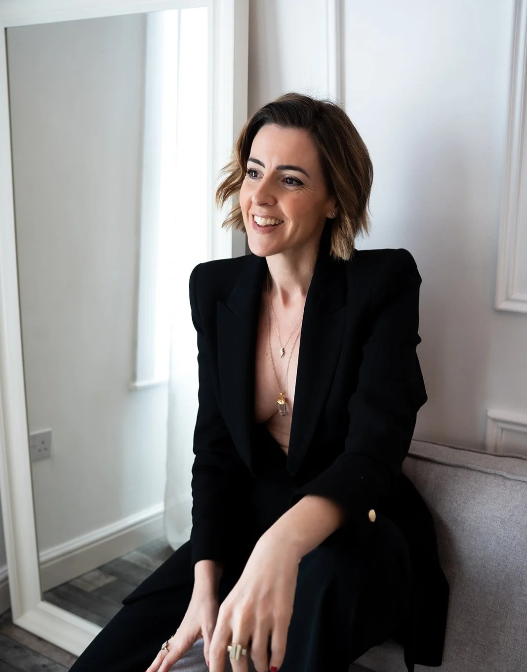 Founder Lisa Moore wearing a black blazer and jewelry, sitting on a light gray sofa near a window with white curtains, smiling and looking to the side.