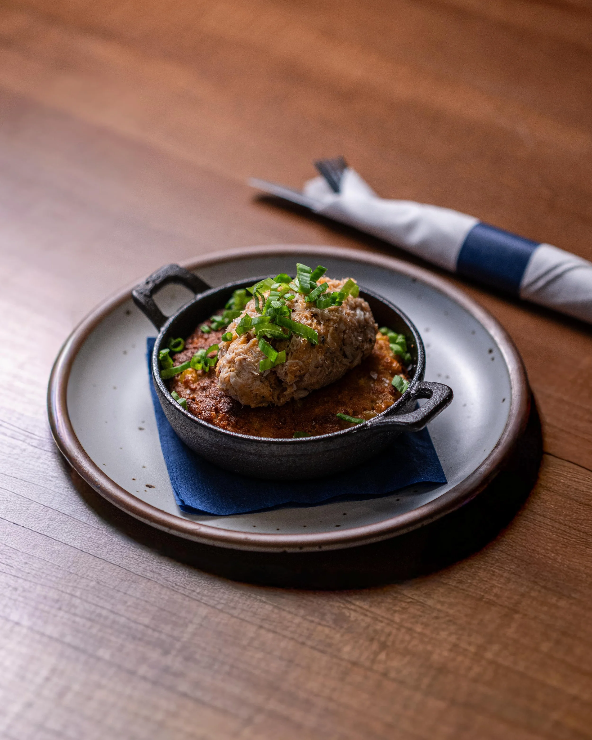 A small black skillet with a cooked meatloaf topped with chopped green onions, served on a white plate with a blue napkin underneath, on a wooden table.