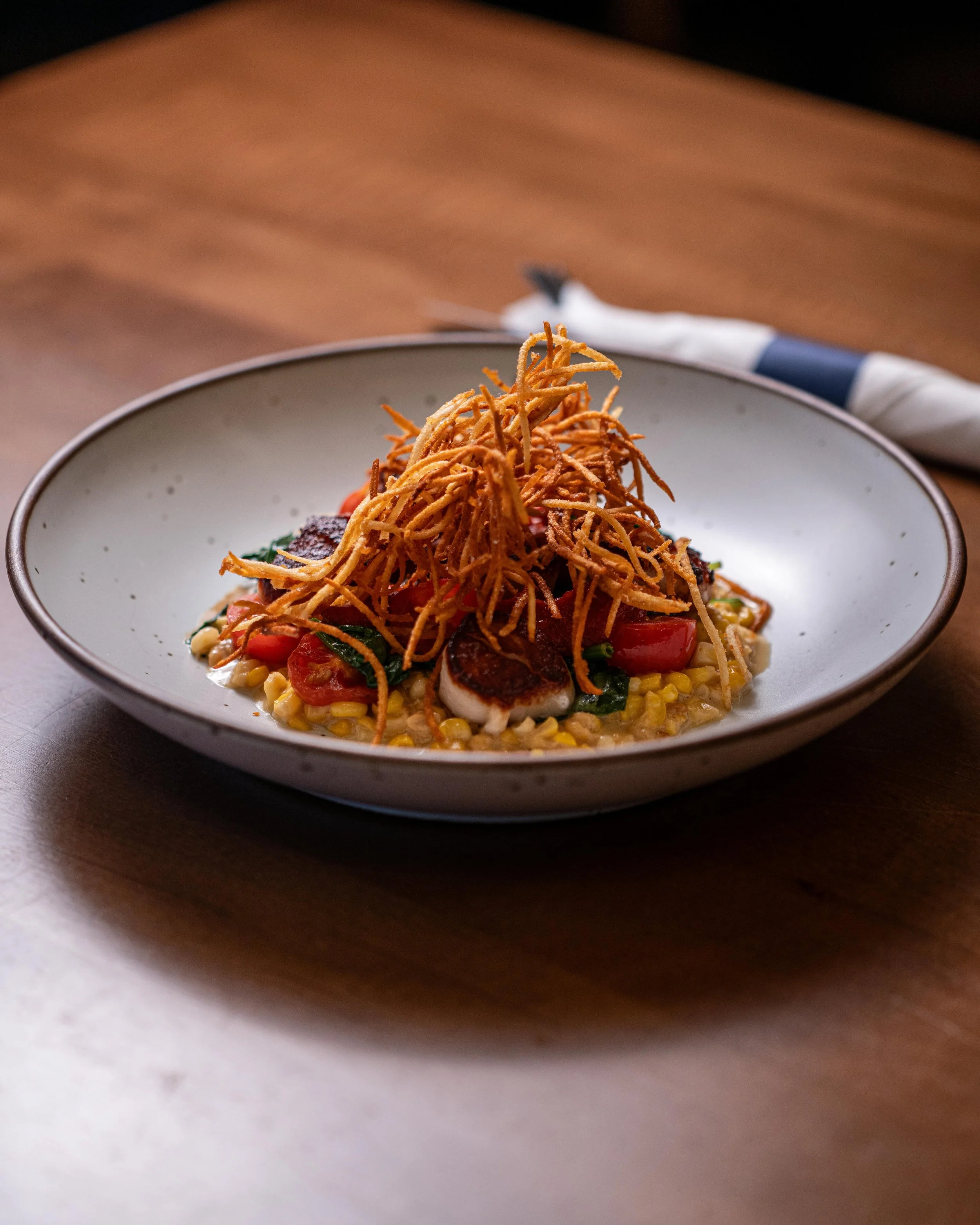 A dish of food served in a shallow bowl with a cream-colored interior and a thin dark rim. The dish includes a bed of yellow corn, topped with slices of grilled meats, cherry tomatoes, and green leafy vegetables, crowned with a crispy, tangled nest o