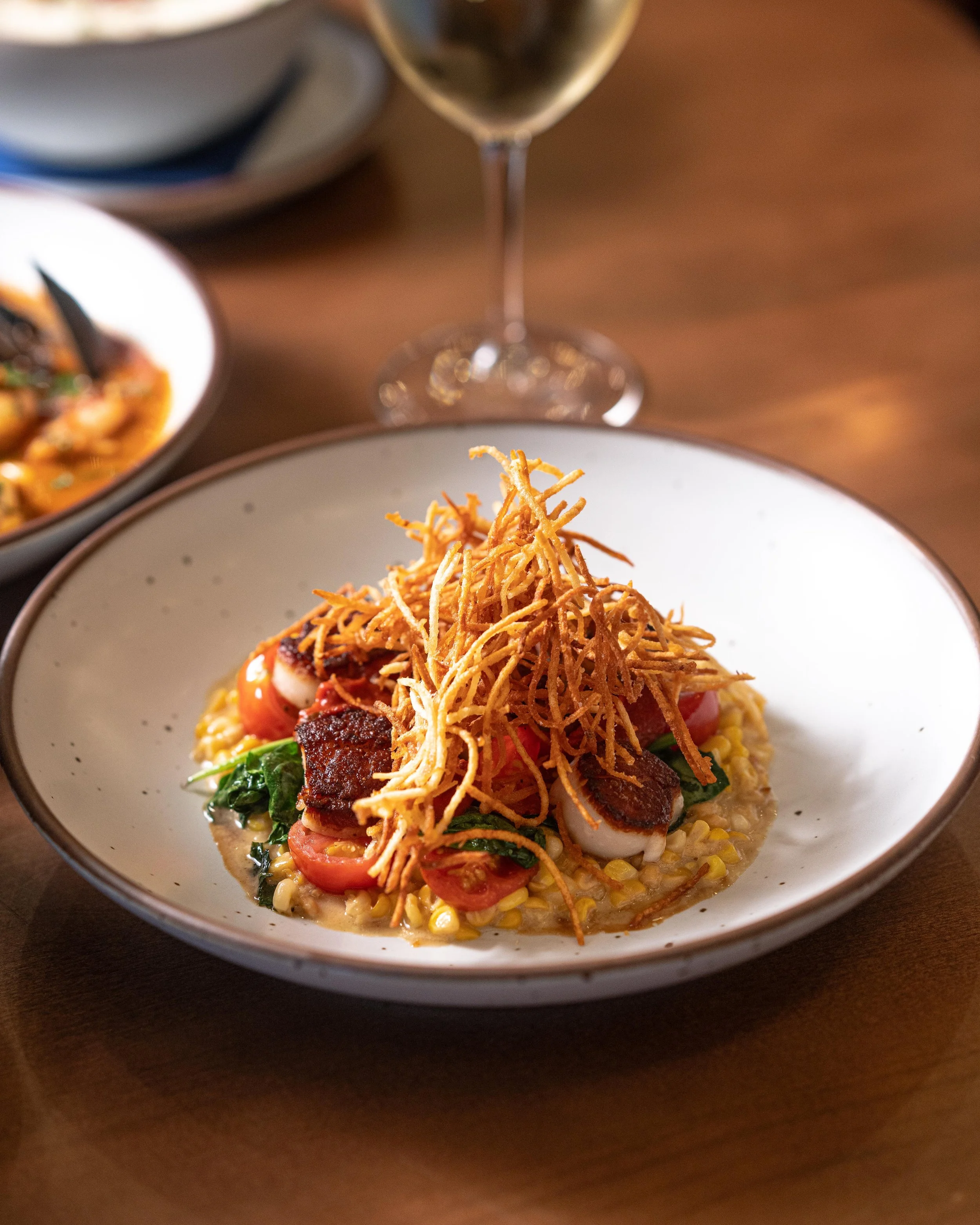 A bowl of gourmet food with a bed of corn grits, topped with cherry tomatoes, wilted greens, crispy shredded fried yuca, and slices of seared meat, served on a wooden table with a glass of red wine and additional dishes in the background.