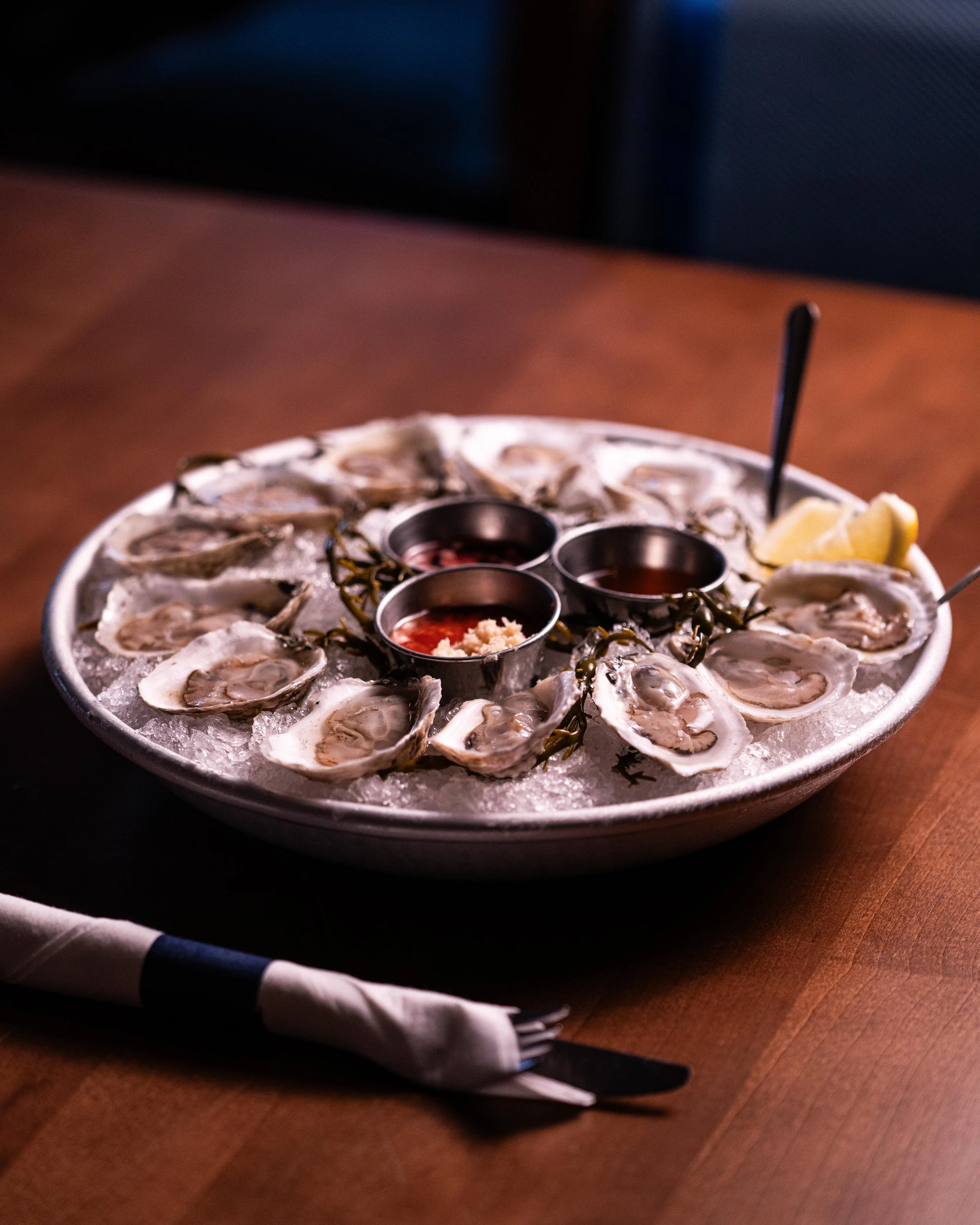 A round seafood platter with oysters on ice, lemon wedges, and dipping sauces, placed on a wooden table with a napkin and utensils nearby.