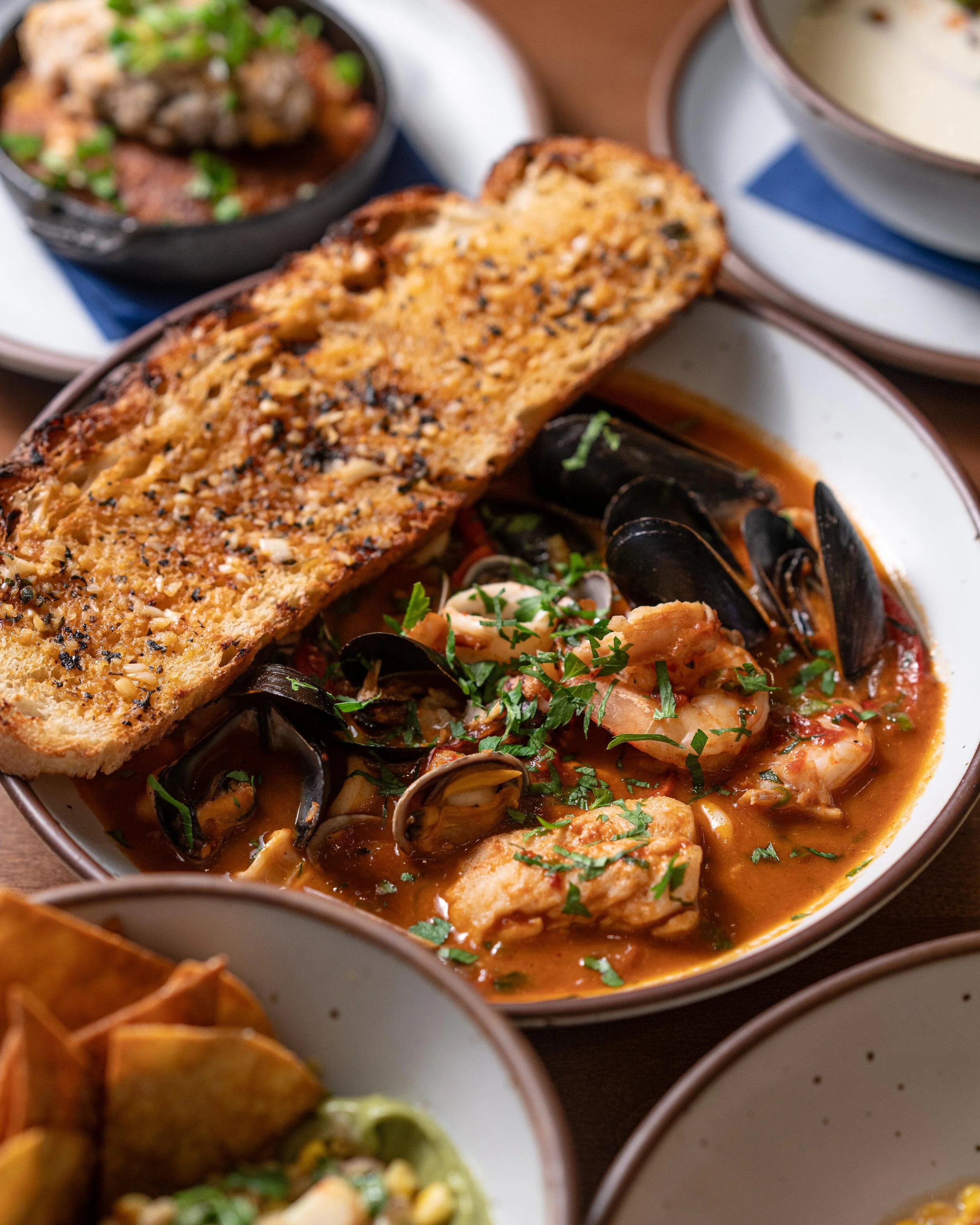 A variety of bowls filled with different types of seafood, including mussels and shrimp, in sauces and garnished with herbs, with slices of garlic bread on the side.