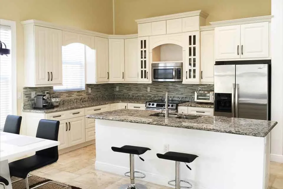 Modern kitchen with white cabinets, granite countertops, stainless steel refrigerator, microwave, oven, and small appliances. Two black bar stools at a breakfast bar, with a window letting in natural light.