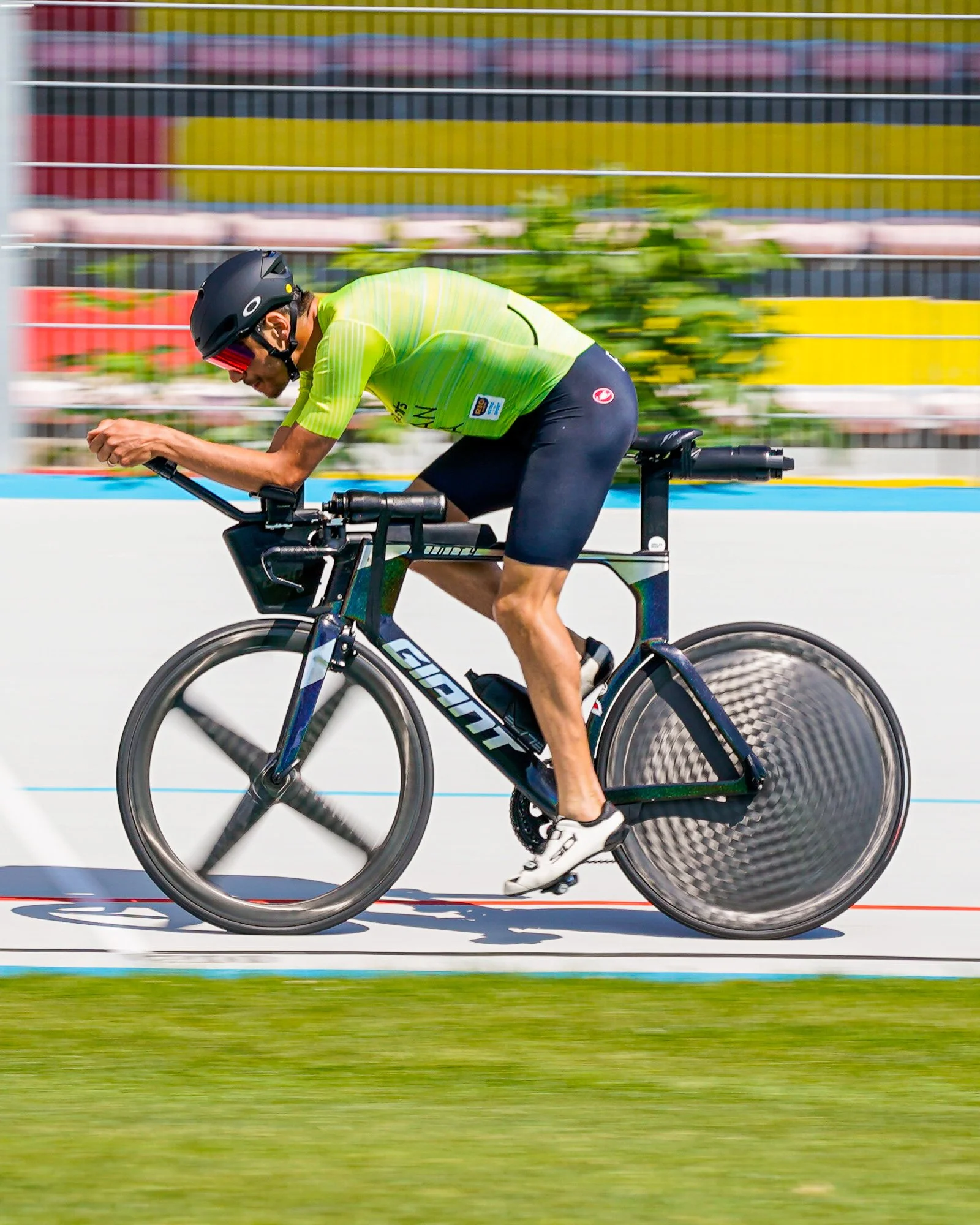 Un uomo in bicicletta professionale in corsa in una pista, indossa un casco nero e una maglietta gialla, con sfondo blurred di ringhiere e vegetazione.
