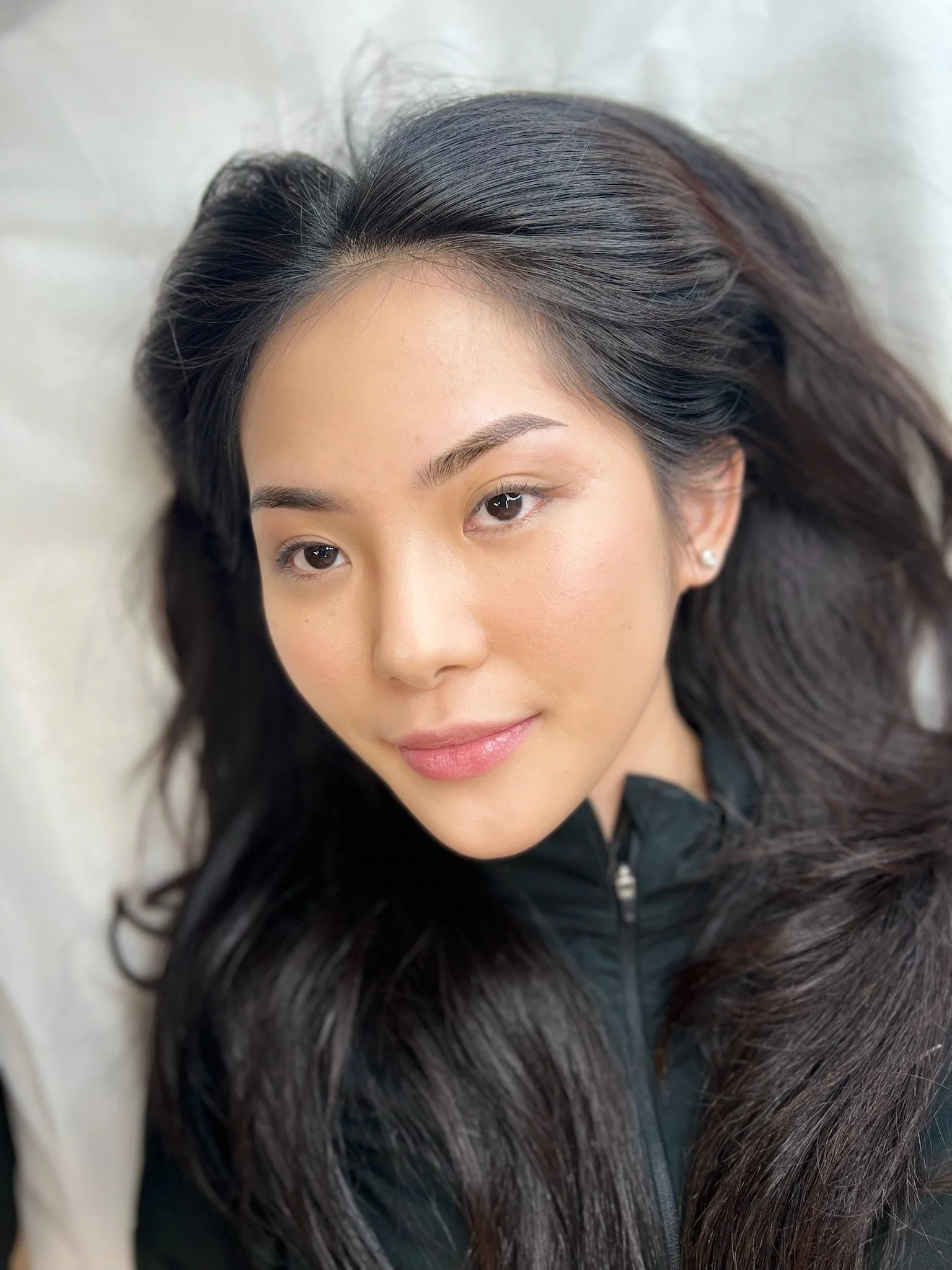 Close-up of a young woman with long dark hair, light makeup, and a black zip-up top, lying on a white surface, smiling softly at the camera.
