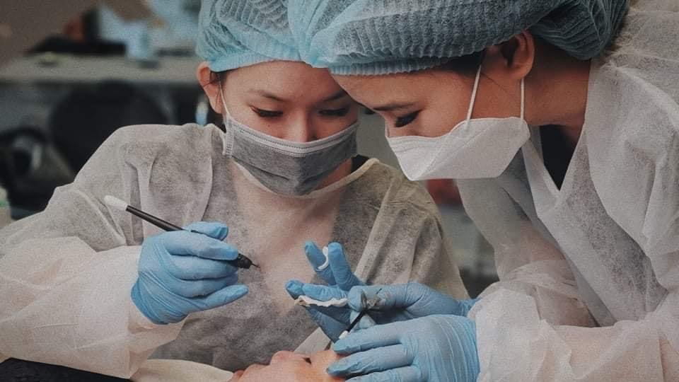 Two healthcare professionals in scrubs, masks, gloves, and hair covers perform a medical procedure together.