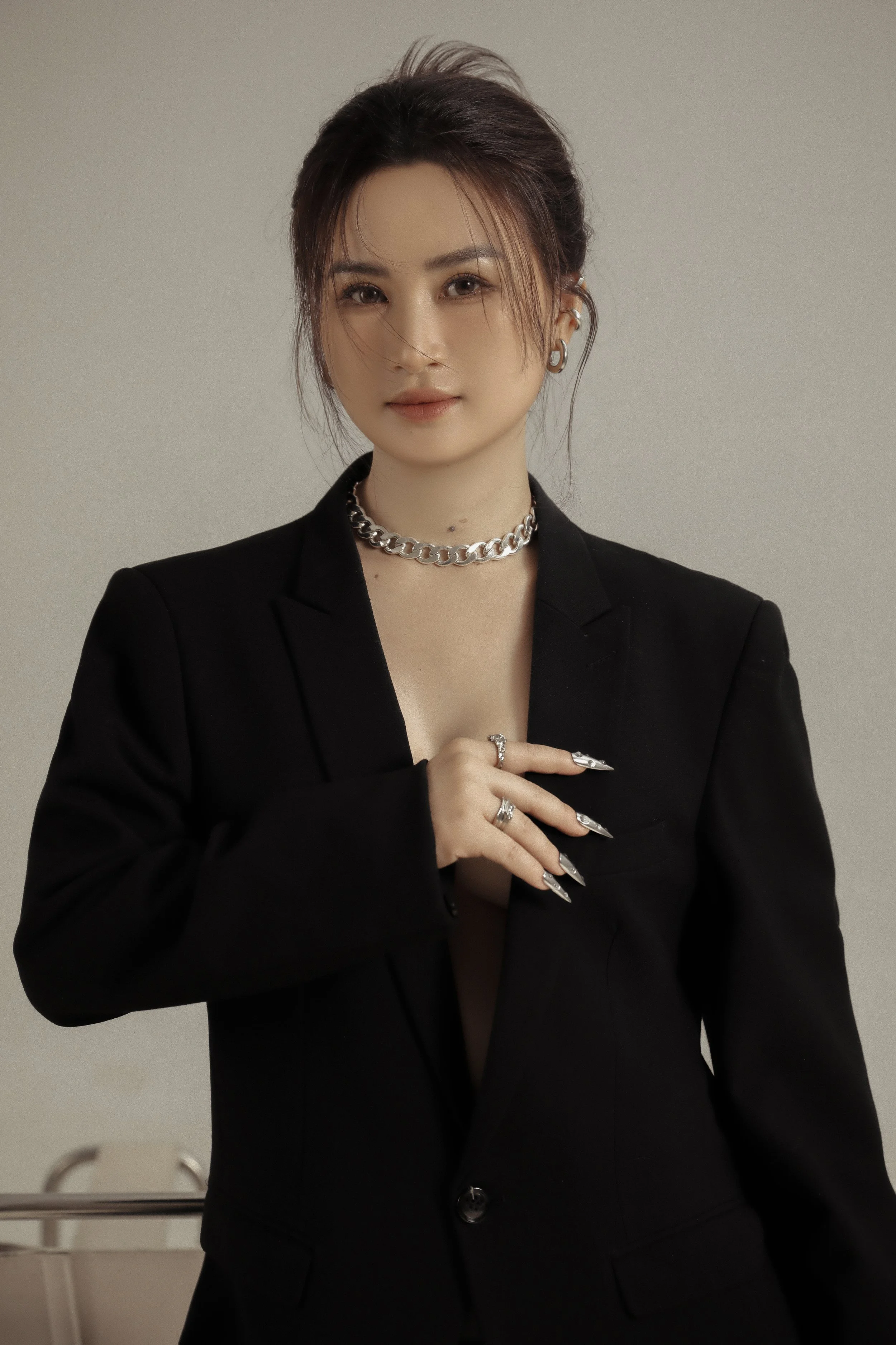 A woman wearing a black blazer with a deep neckline, accessorized with silver jewelry, including a chain necklace and multiple rings, posing against a plain background.