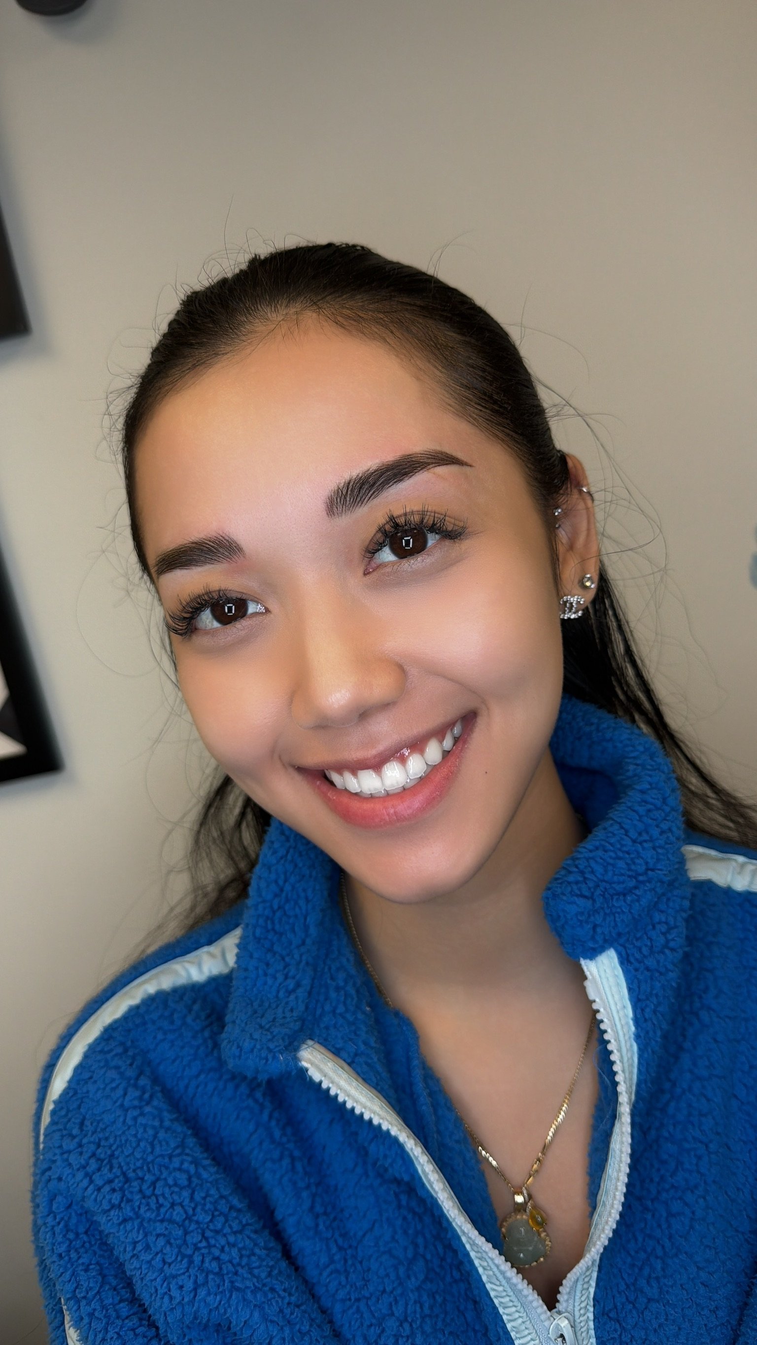A young woman with dark hair, smiling, wearing earrings and a blue zip-up fleece jacket, indoors.