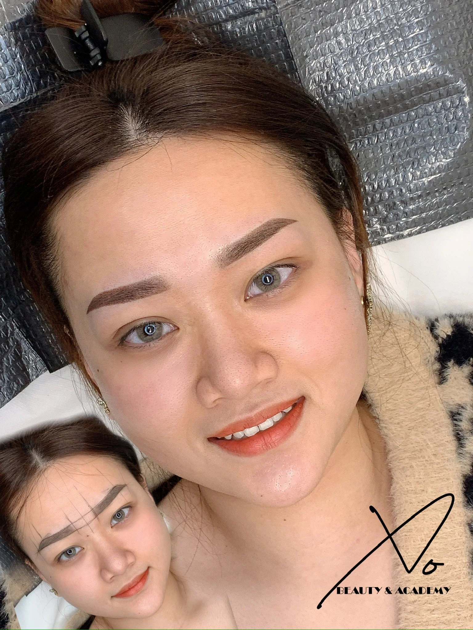 A woman with well-groomed eyebrows, light makeup, and a leopard-print top lying on a beauty salon bed, with another woman with drawn eyebrows in the background, both looking at the camera, with a logo reading 'Beauty & Academy' at the bottom right.