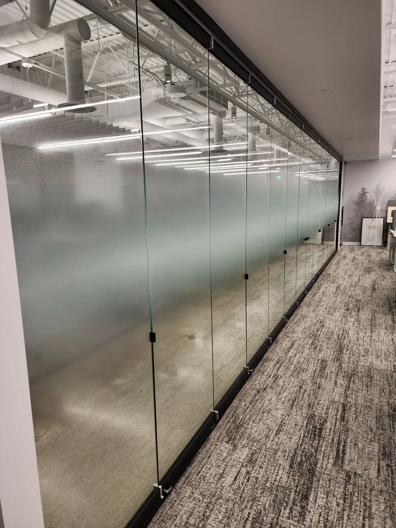 Glass partition with frosted middle section and clear top and bottom sections, in an office setting with visible ceiling ducts and lighting.