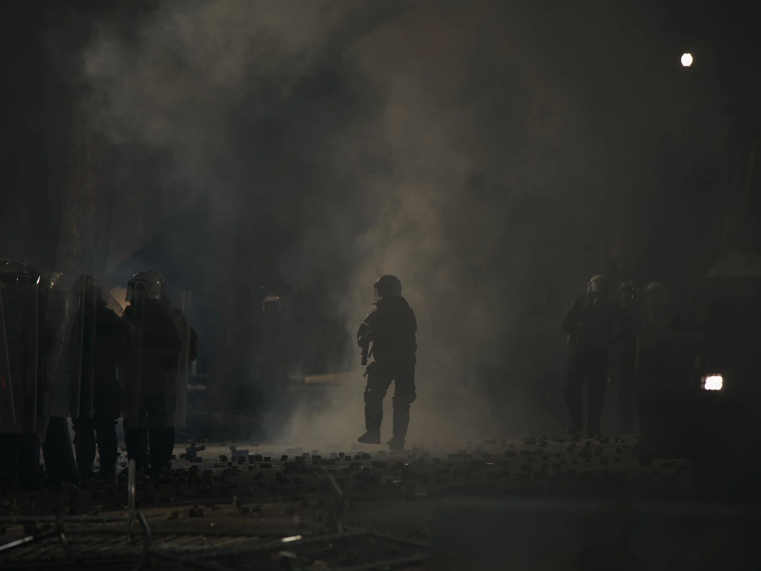 A police officer walks through tear gas and stones thrown by demonstrators, holding a tear gas launcher ready to fire.