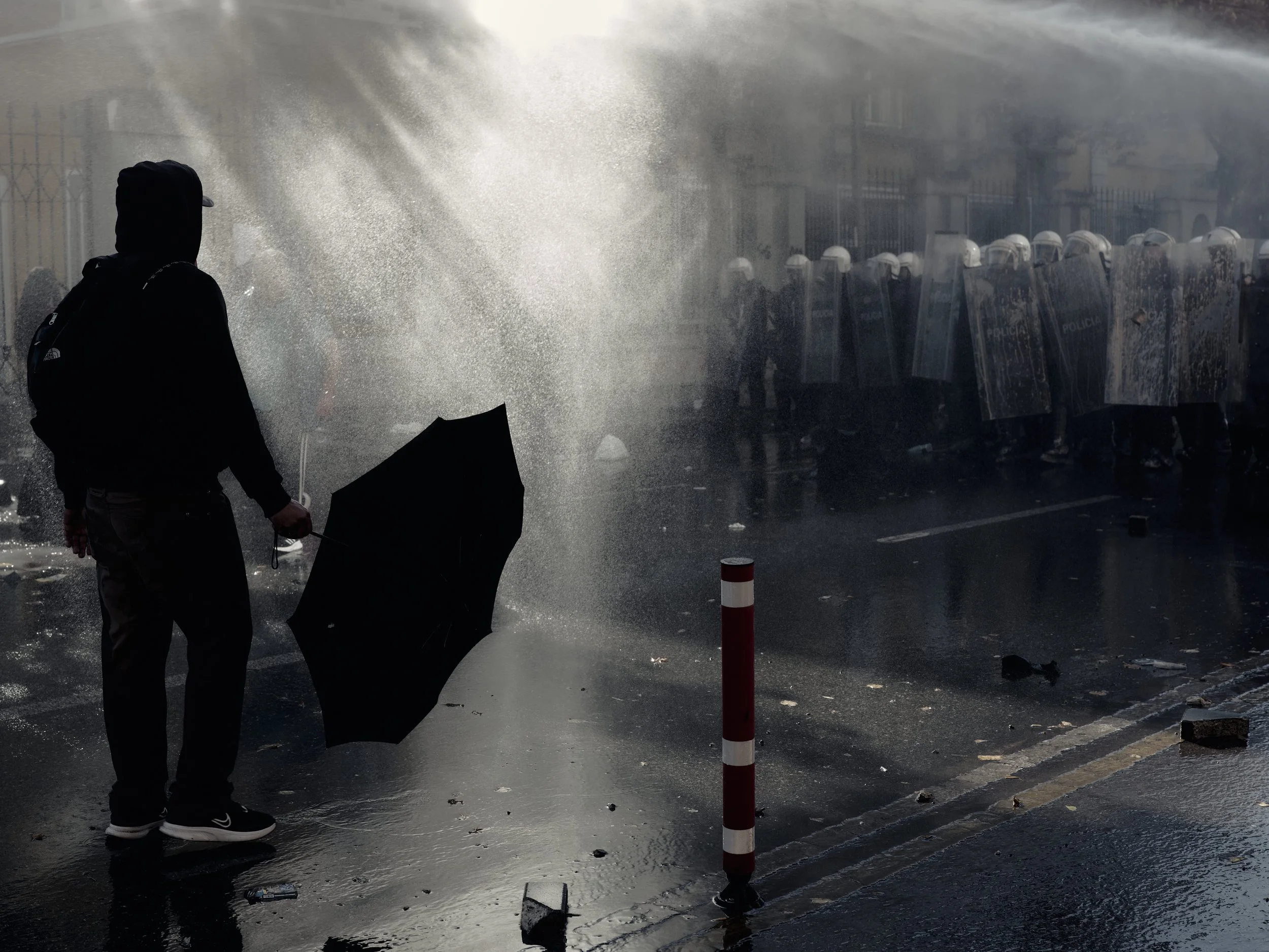 A student stands in front of police holding an umbrella as water sprays overhead.