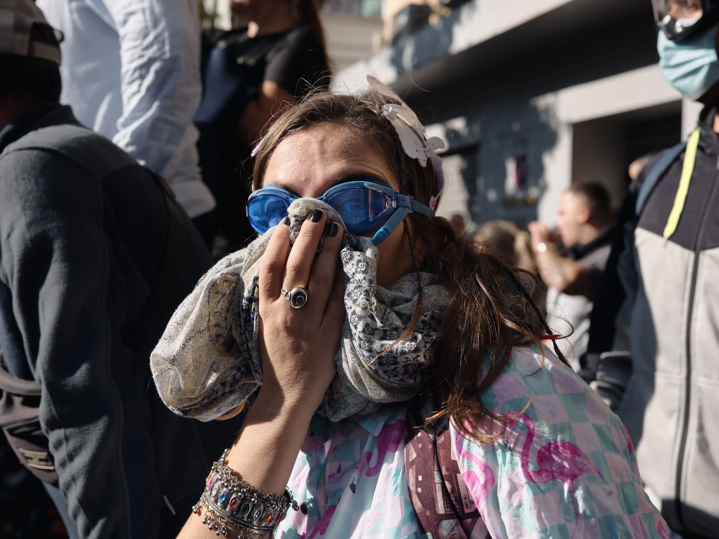 A woman wearing a T-shirt printed with flamingos and protective glasses shields her face from tear gas, holding her scarf over her nose and mouth.