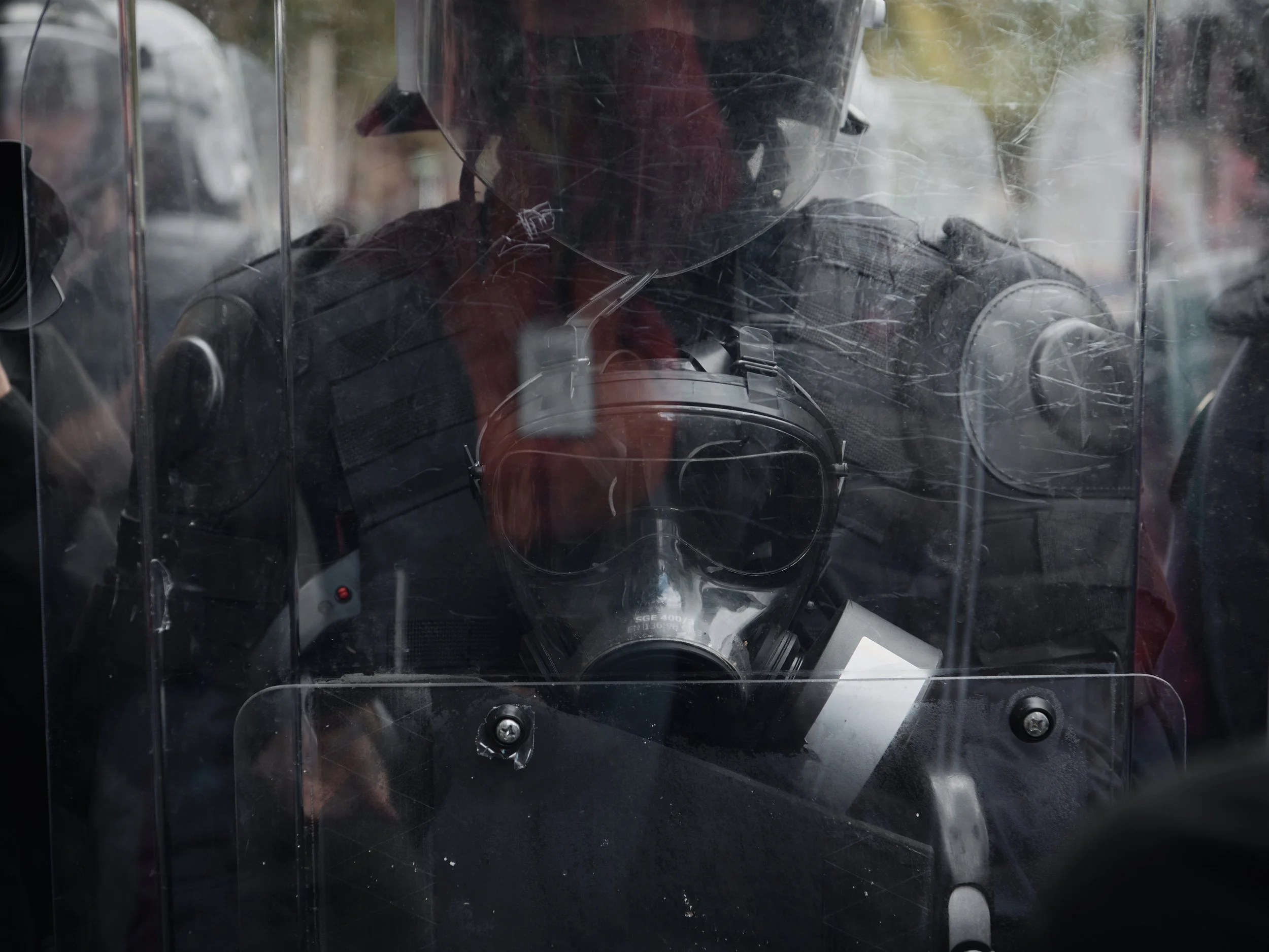 A police officer’s face is partially visible behind a shield moments before officers begin dispersing demonstrators near the Albanian Parliament.