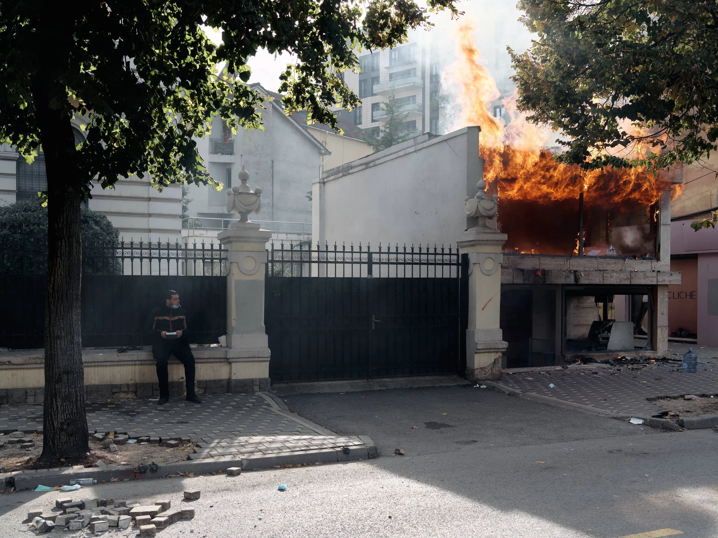 A shop burns at the beginning of Rruga e Durrësit as a man sits nearby, smoking a cigarette.