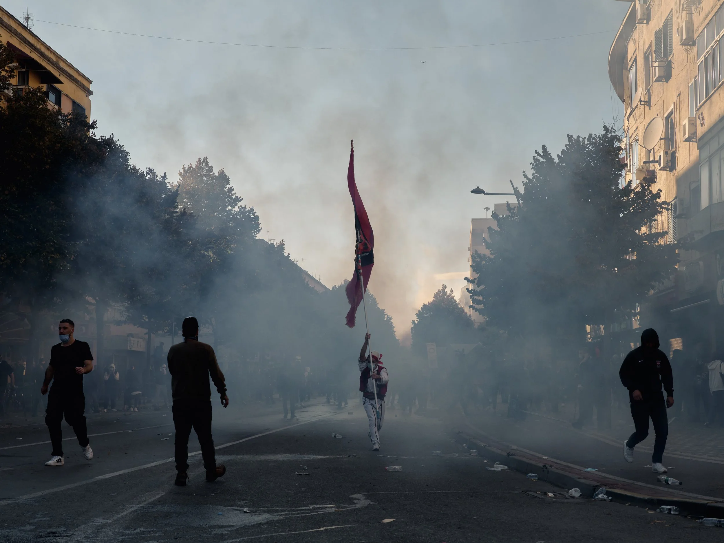 A man wearing traditional Albanian clothing and carrying the Albanian flag walks away from tear gas on Rruga e Durrësit.
