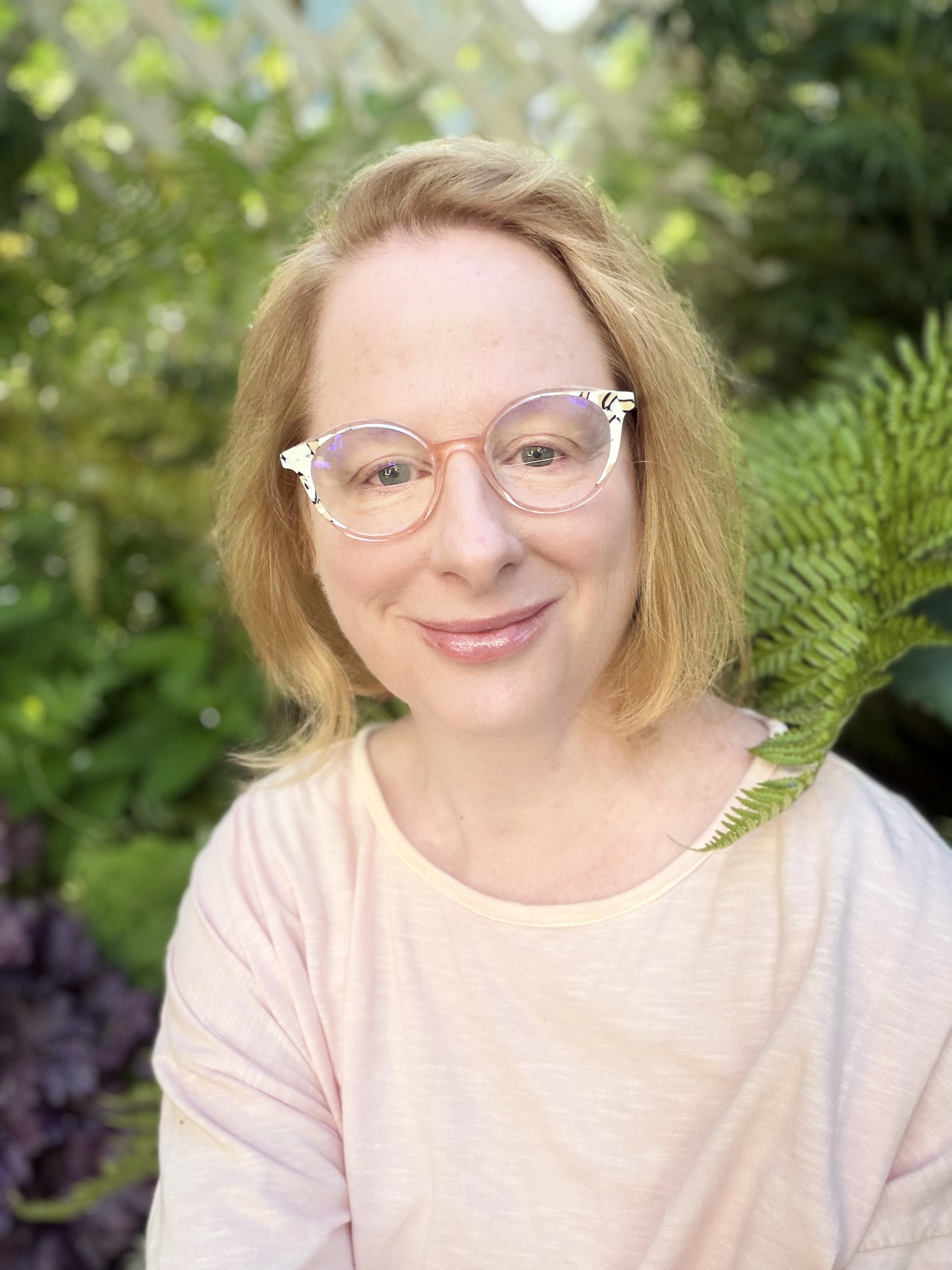 A woman with blonde hair and glasses smiling outdoors surrounded by green foliage.