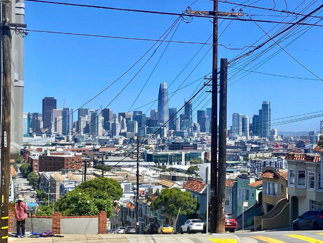 A view of San Francisco skyline with skyscrapers in the distance, viewed from a neighborhood street with houses and trees in the foreground, utility poles, and power lines.