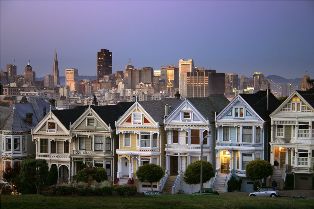 A row of colorful Victorian-style houses with the San Francisco skyline in the background during dusk.