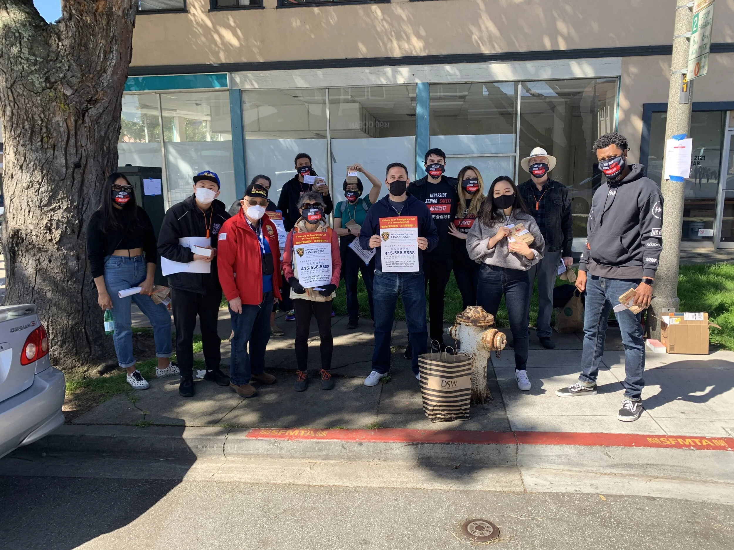 A group of people wearing face masks standing on a sidewalk in front of a building, some holding flyers and boxes, participating in a community event or distribution.