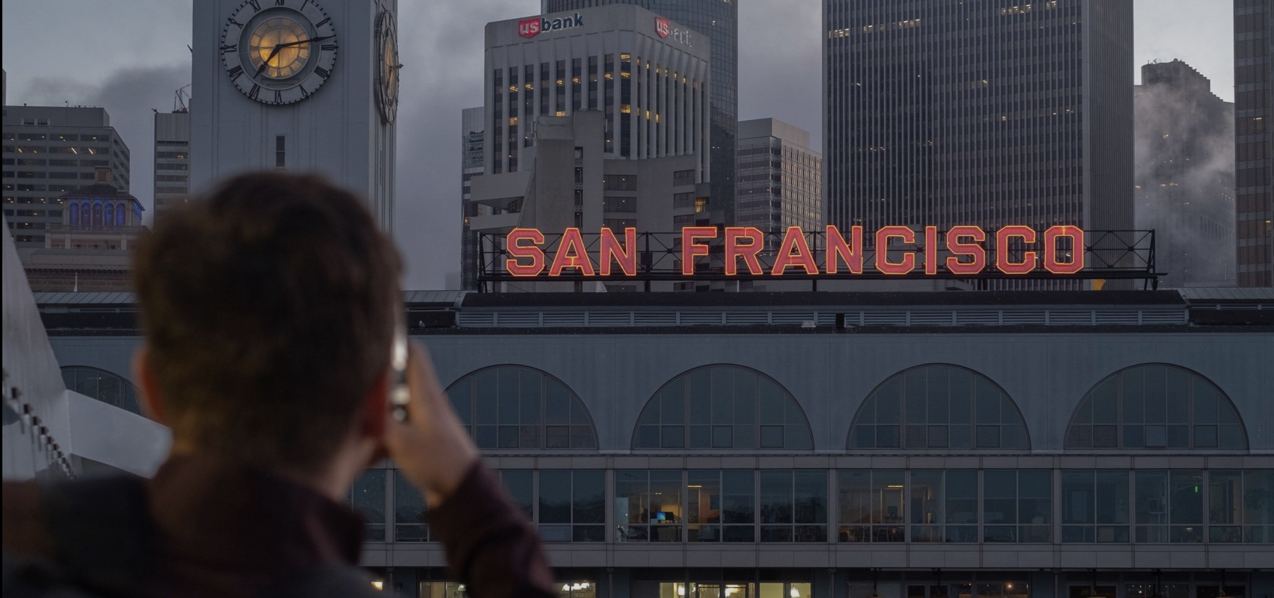 Person taking photo of San Francisco skyline with neon sign, skyscrapers, clock tower, and cloudy sky.
