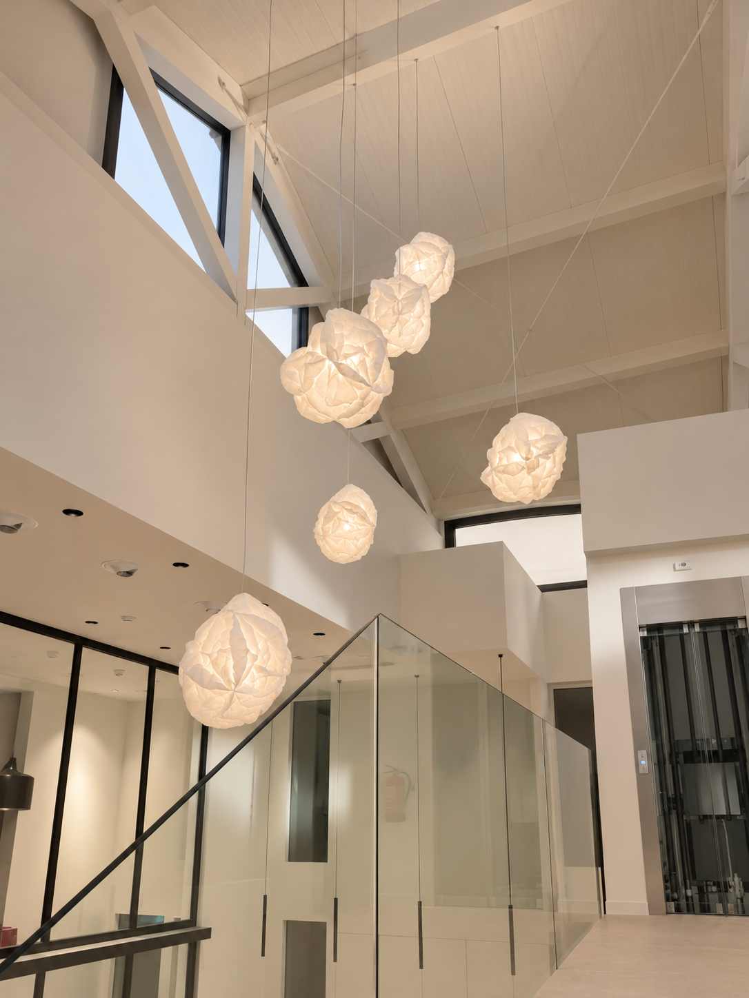 Racimo de lámparas de papel plegado en forma de nube suspendidas sobre una escalera con barandilla de cristal — solución de iluminación ZONA