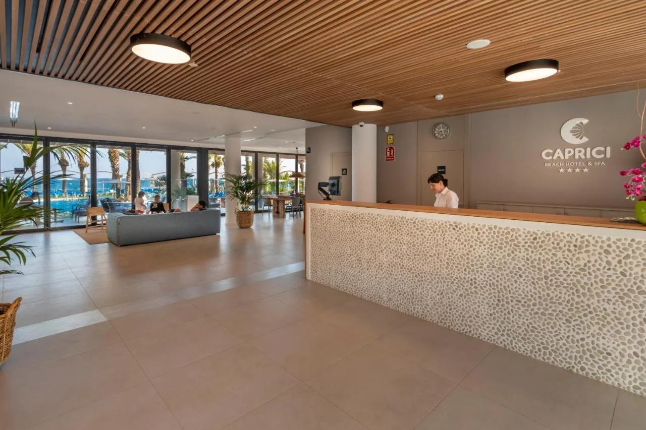 Recepción de Caprici Beach Hotel & Spa con mostrador revestido en piedra y vistas a la piscina y el mar