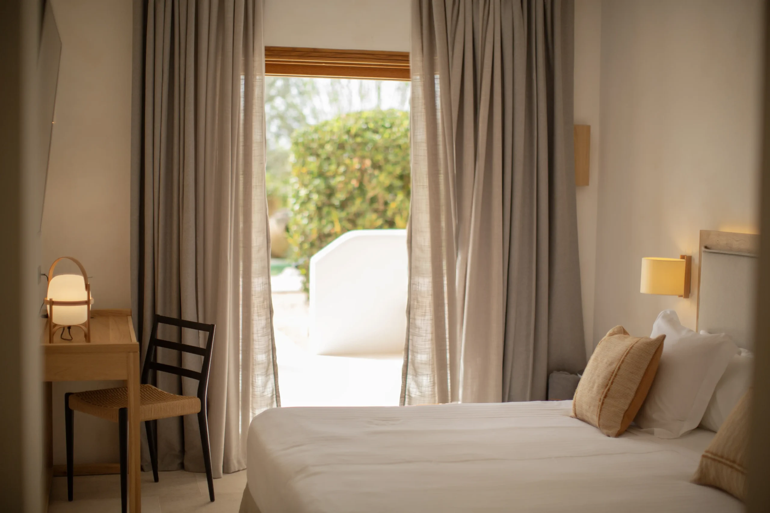 Habitación de Can Jaume con cortinas de lino, escritorio de madera y puerta acristalada al jardín