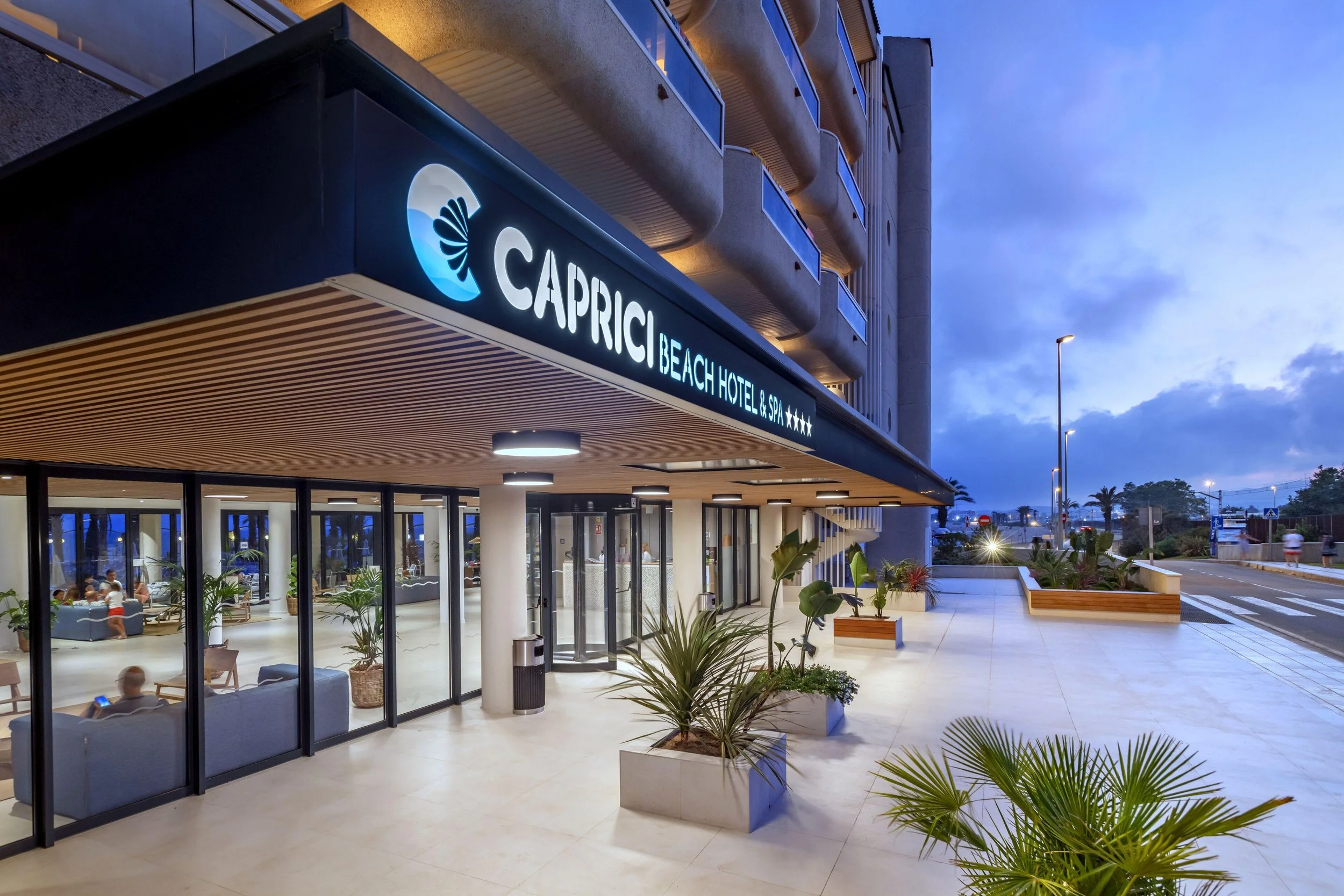 Entrada de Caprici Beach Hotel & Spa al anochecer, con letrero iluminado y fachada renovada