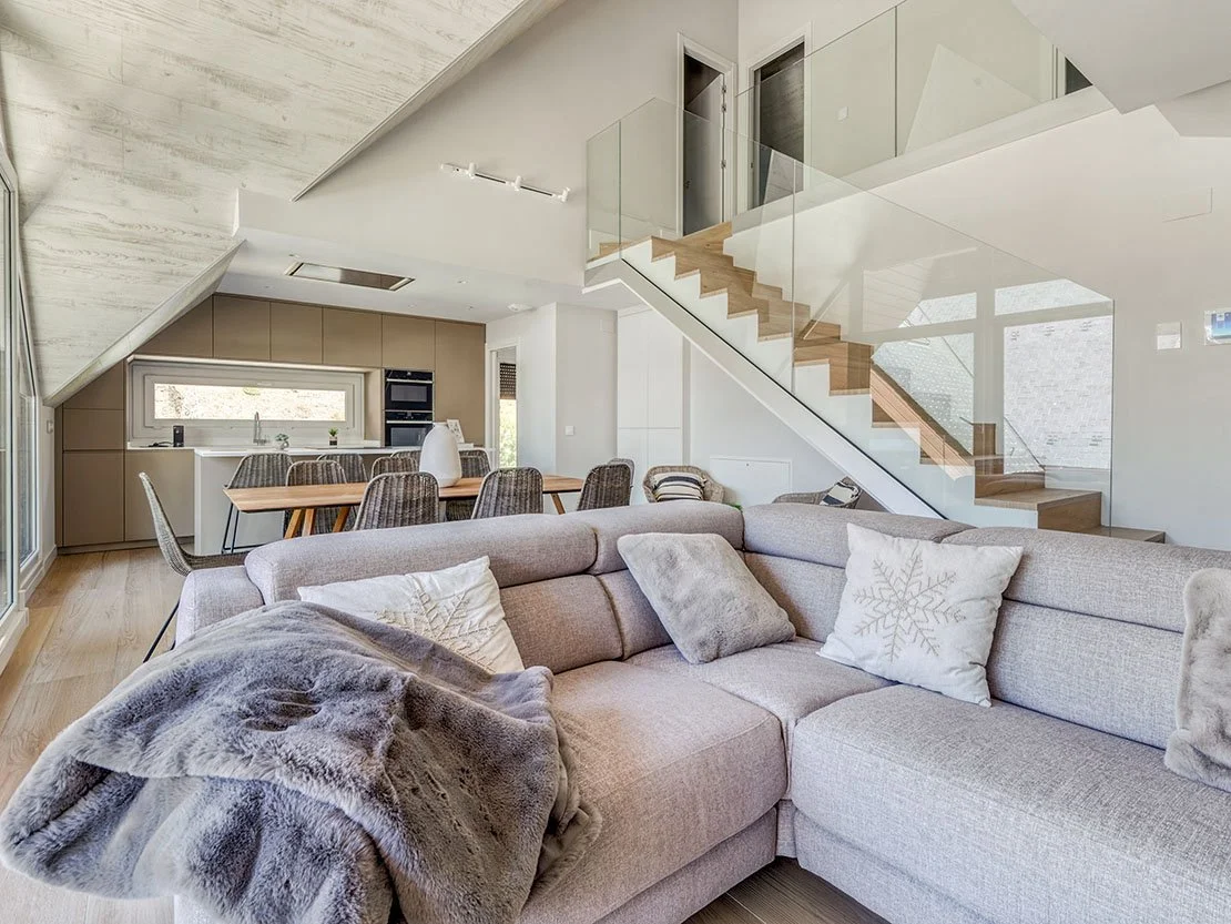 Salón de Residencial Els Pletieus con sofá de esquina, mantas de invierno y escalera de cristal hacia la planta superior