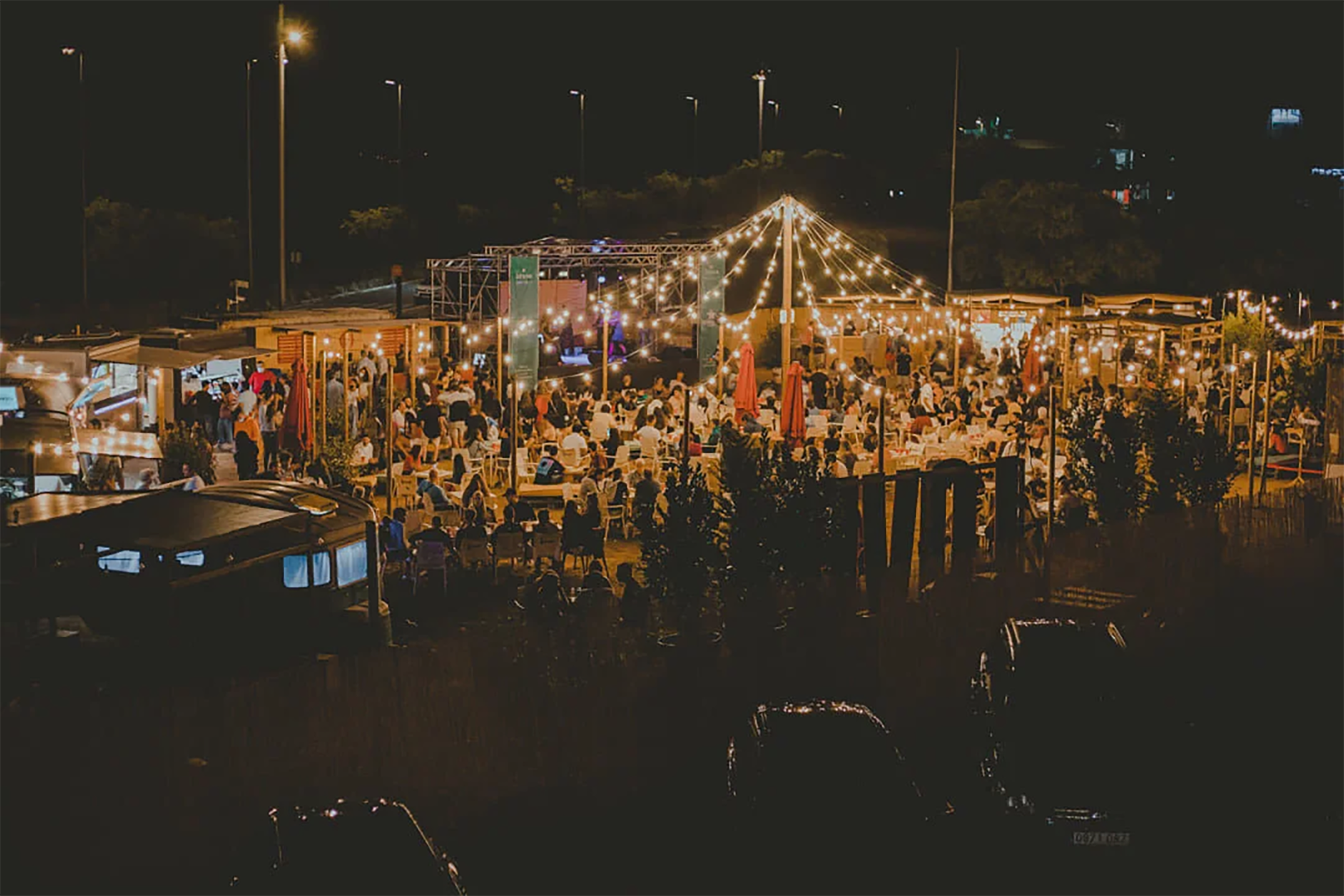 Vista aérea nocturna de Itaca Market con escenario, público y guirnaldas de luces sobre las mesas
