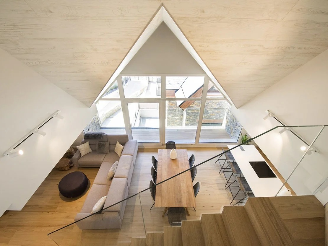 Vista elevada del salón, comedor y cocina de Val d'Aran, con gran ventanal triangular y estructura de madera vista