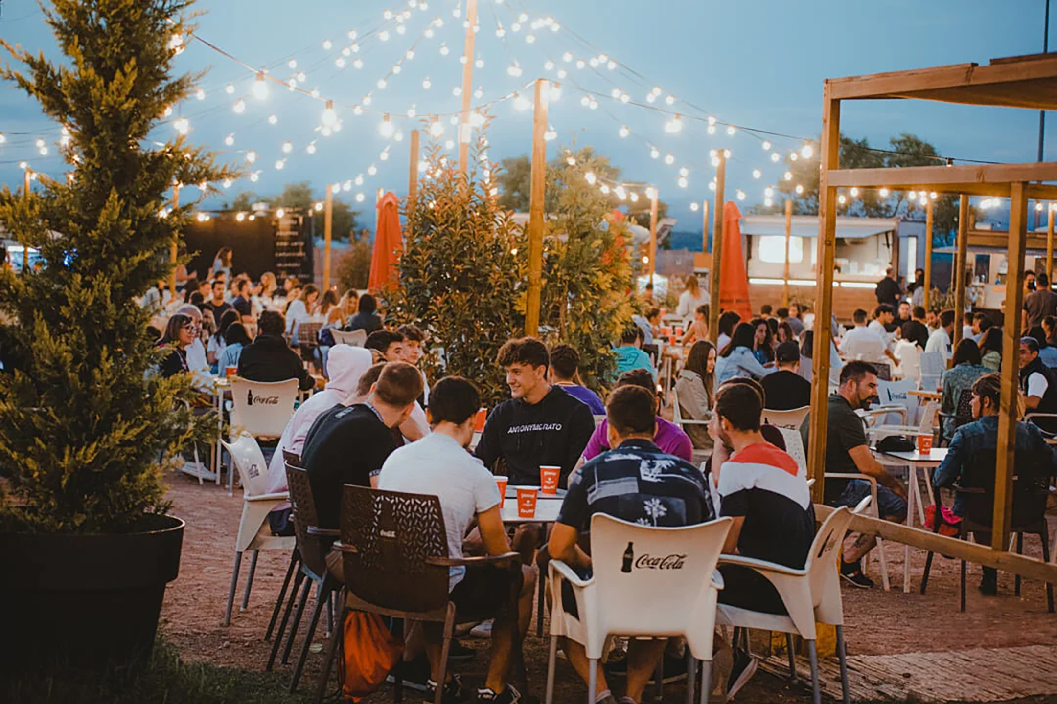 Ambiente nocturno en Itaca Market, con mesas al aire libre, guirnaldas de luces y food truck al fondo