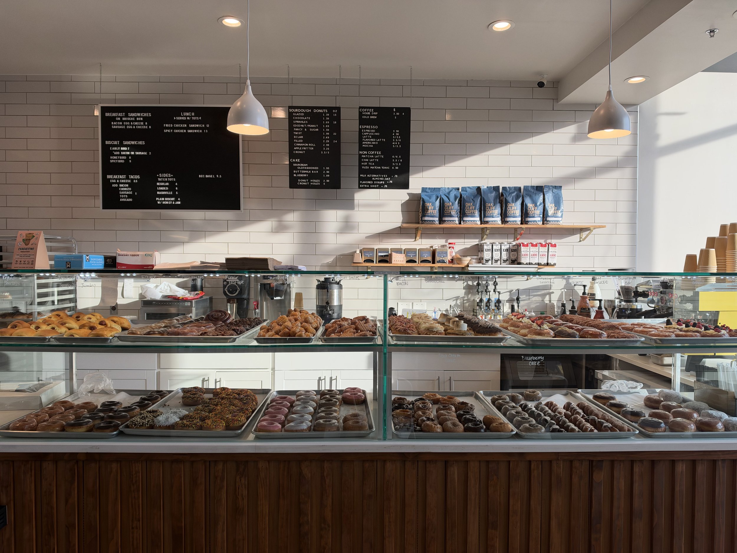 Display case at a bakery filled with various donuts and baked goods, with menu boards and coffee supplies on the wall behind.