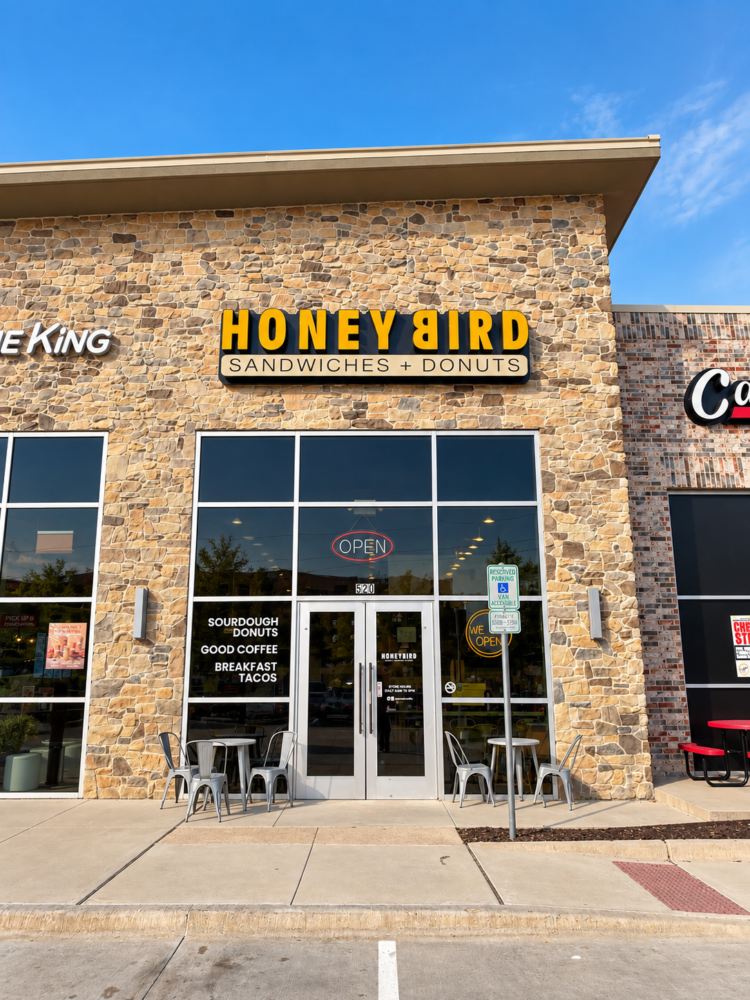 Exterior of Honeybird restaurant with stone facade, large glass windows, seating outside, and a sign that reads 'HONEYBIRD SANDWICHES + DONUTS.'