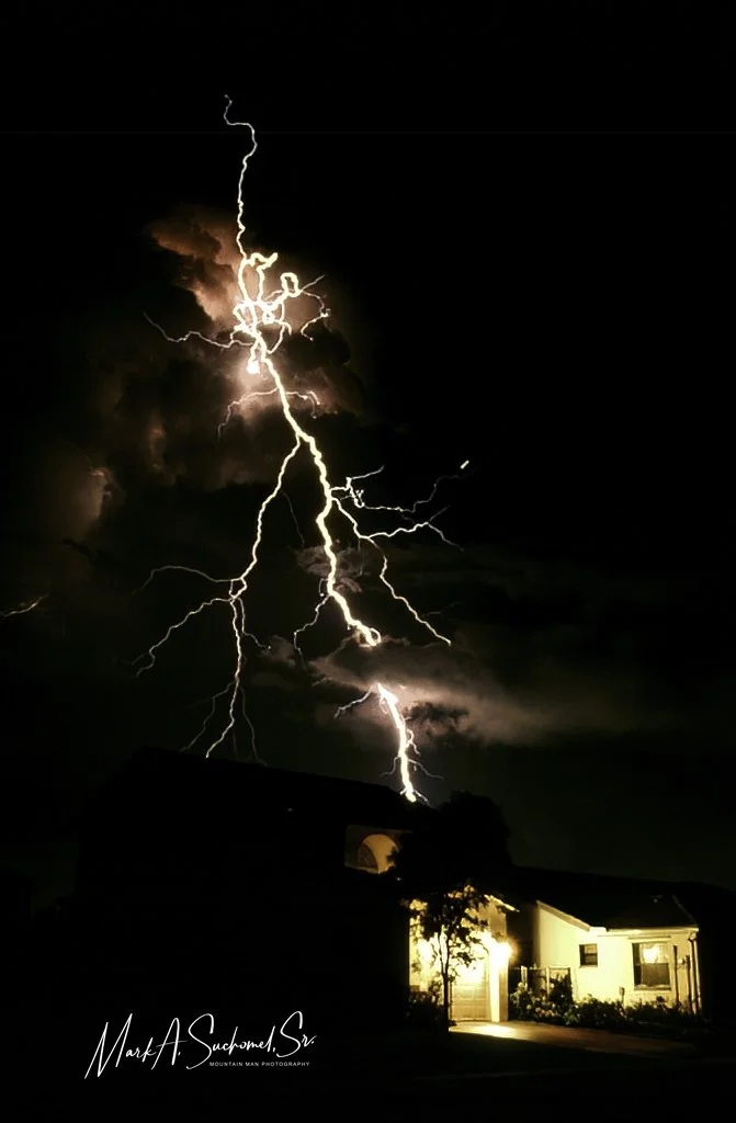 Lightning strikes during a thunderstorm at night, illuminating dark clouds and a residential house with a lit garage and trees in the foreground.